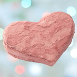 The Liberator Heart Wedge Fur - Rose, a pink, heart-shaped plush pillow from Liberator with a soft textured surface, is displayed against a blurred background of pastel lights.