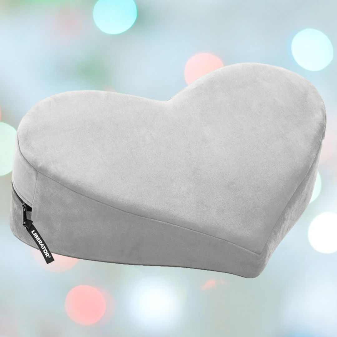 The Liberator Heart Wedge MicroVelvet - Grey by Liberator is shown against pastel lights, featuring a heart shape, soft microvelvet cover, and side zipper—ideal as a cozy derriere positioning aid or stylish home decor accent.
