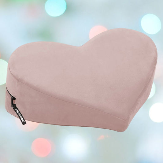 The Liberator Heart Wedge MicroVelvet - Rose, a light pink heart-shaped pillow by Liberator with a side zipper tag, is displayed against a softly blurred background of pastel blue, green, and pink circles. 1080