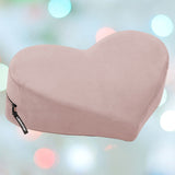 The Liberator Heart Wedge MicroVelvet - Rose, a light pink heart-shaped pillow by Liberator with a side zipper tag, is displayed against a softly blurred background of pastel blue, green, and pink circles.