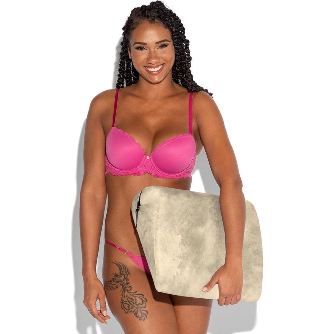 A smiling woman with long curly hair, wearing a pink bra and matching underwear, holds a beige zippered pouch—ideal for storing essentials like the Liberator Jaz Ibiza Collection - Natural sex position pillow by Liberator. Her left thigh shows a floral tattoo.
