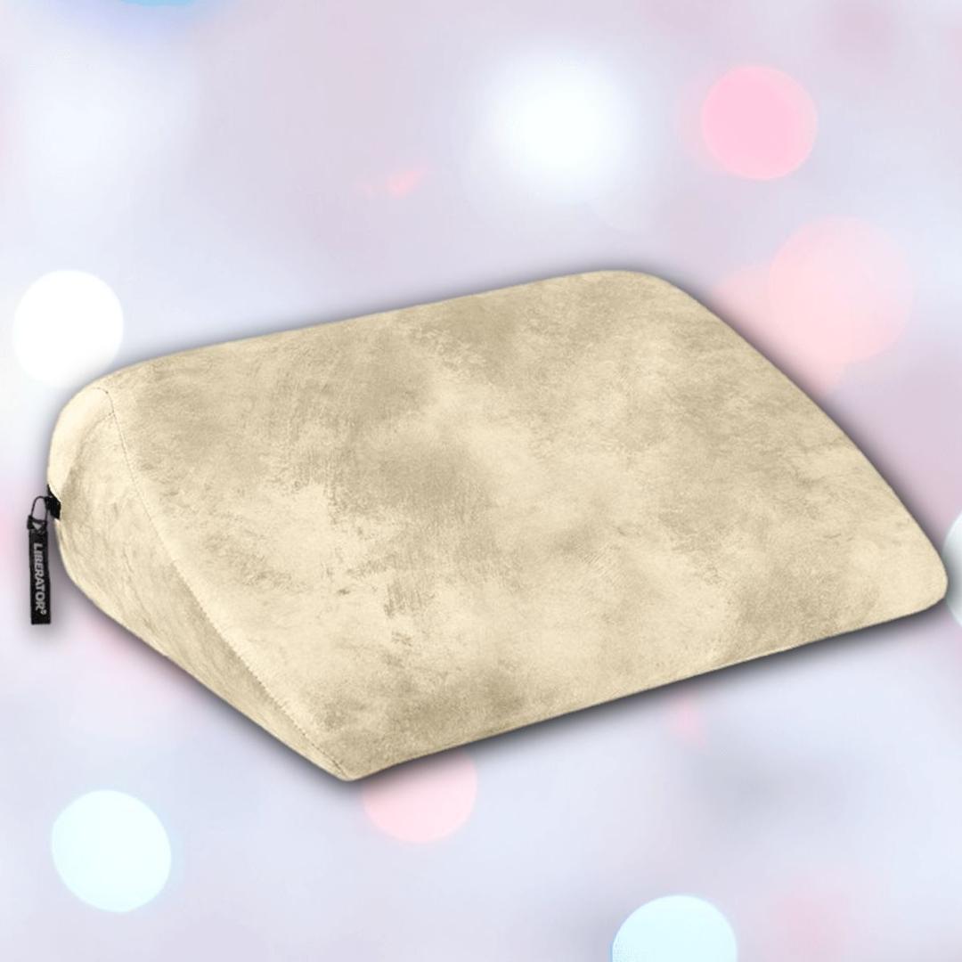 A Liberator Jaz Ibiza Collection - Natural pillow by Liberator sits on a soft-focus pastel background with pink, blue, and white light spots. Its smooth beige surface is ideal for G-spot stimulation and features a small black tag on the side.