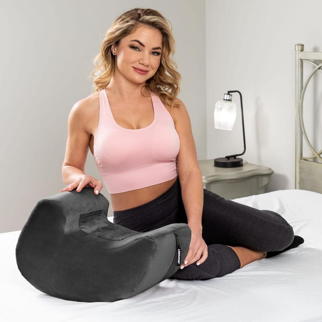 A woman wearing a pink sports bra and dark leggings sits on a bed, smiling with one arm resting on the Liberator Pulse MicroFiber - Black by Liberator. A small lamp and bedside table can be seen in the background.