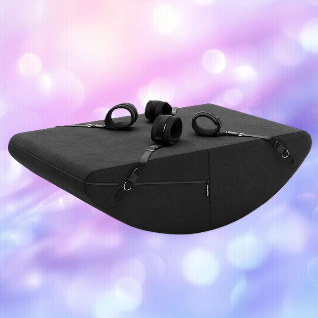 The Liberator Scoop Rocker with Microloop & Cuffs in black by Liberator is a wedge-shaped cushion with built-in cuffs and adjustable straps, shown against a softly blurred background of pastel pink, purple, and blue tones.