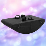 The Liberator Scoop Rocker with Microloop & Cuffs in black by Liberator is a wedge-shaped cushion with built-in cuffs and adjustable straps, shown against a softly blurred background of pastel pink, purple, and blue tones.