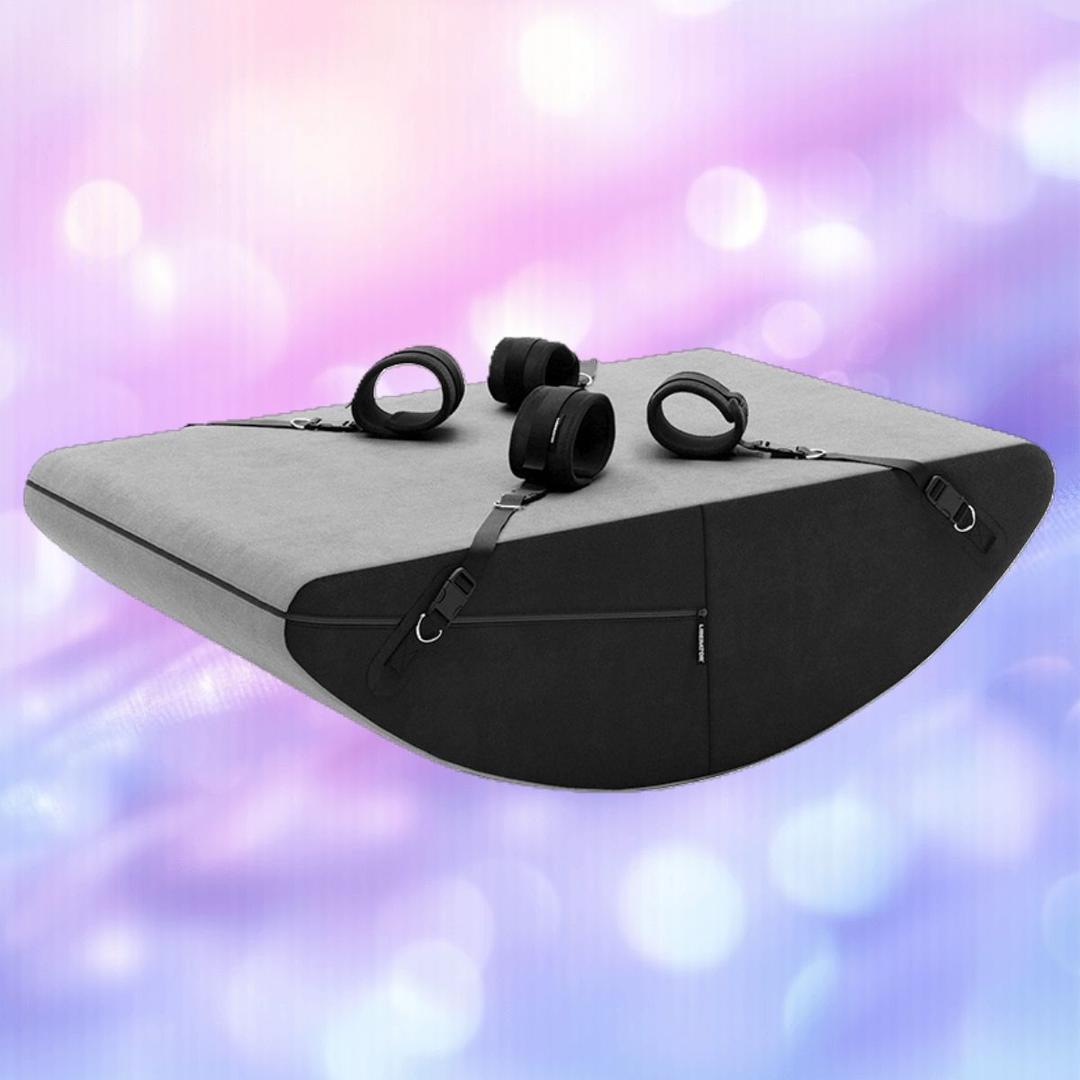 The Liberator Scoop Rocker: MicroLoop Bondage Edition with Cuffs in grey by Liberator, featuring a wedge shape and four adjustable black straps, is displayed against a blurred pink and purple background.