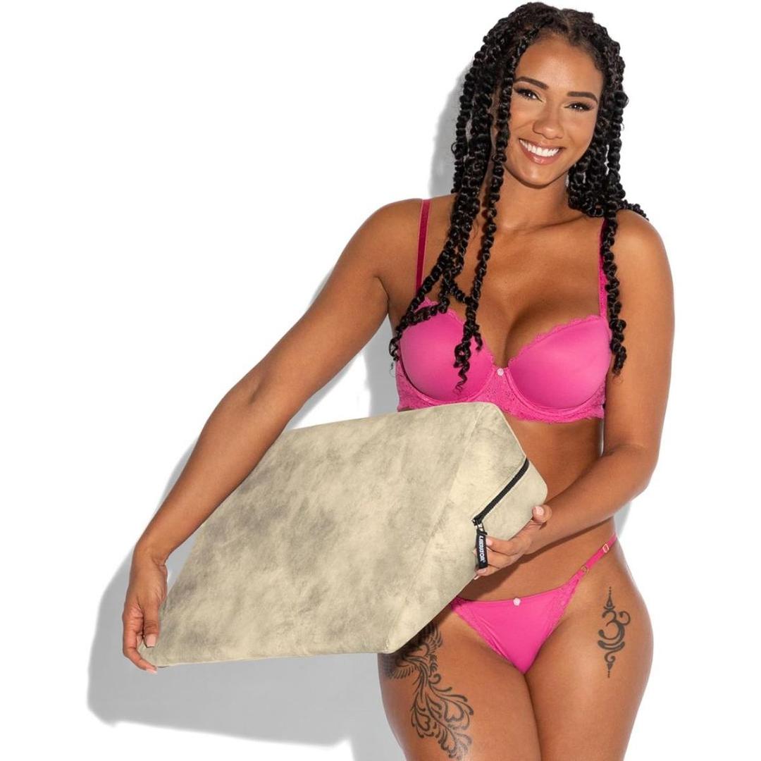 A woman with long, curly hair and floral thigh tattoos smiles while holding the Liberator Wedge Ibiza Collection - Natural by Liberator in front of her. She wears a bright pink lingerie set against a white background.