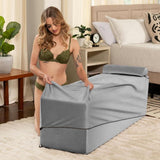 A woman in green lingerie kneels on a beige rug, fitting the Liberator Aria Chaise - Grey by Liberator with its cover. The bedroom features a bed, plants, and light curtains, enhancing a discreet love lounge ambiance.