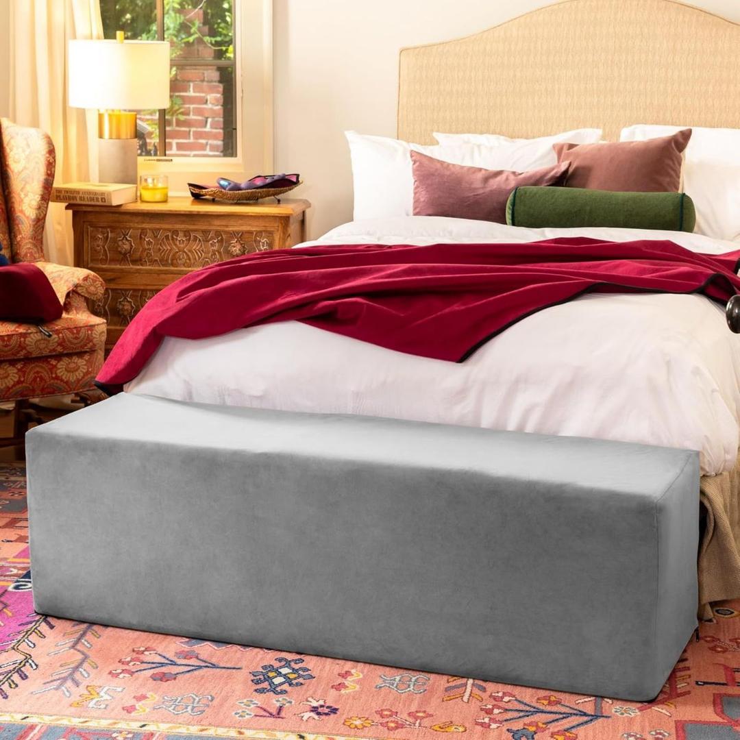 A cozy bedroom features a large bed with white bedding, a red throw, and a patterned rug. At the foot of the bed is the Liberator Aria Chaise in grey by Liberator, while a wooden nightstand with a lamp sits beside the bed.