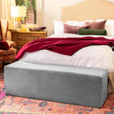 A cozy bedroom features a large bed with white bedding, a red throw, and a patterned rug. At the foot of the bed is the Liberator Aria Chaise in grey by Liberator, while a wooden nightstand with a lamp sits beside the bed.