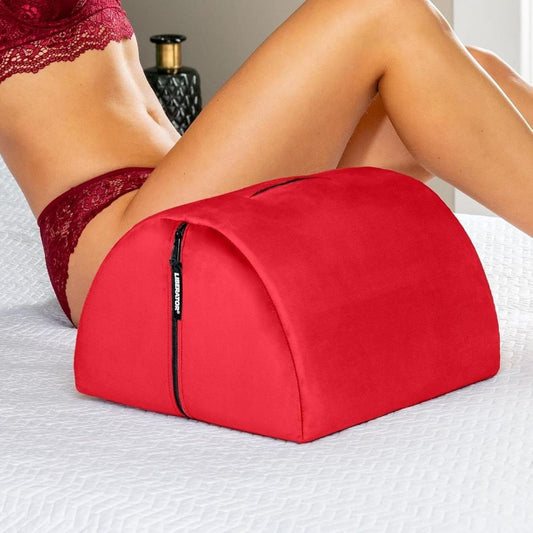 A woman in red lace lingerie leans on the Liberator Bonbon Microvelvet - Flame by Liberator, a red wedge-shaped sex toy mount with a zipper, placed on a white quilted bedspread. The pillow is designed for support and hands-free solo play. 1080