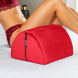 A woman in red lace lingerie leans on the Liberator Bonbon Microvelvet - Flame by Liberator, a red wedge-shaped sex toy mount with a zipper, placed on a white quilted bedspread. The pillow is designed for support and hands-free solo play.