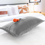 A Liberator Humphrey MicroVelvet - Grey pillow by Liberator with a toy pocket holds an orange and red wand-shaped device, resting on a neatly made bed with decorative pillows and a bedside lamp in the background.