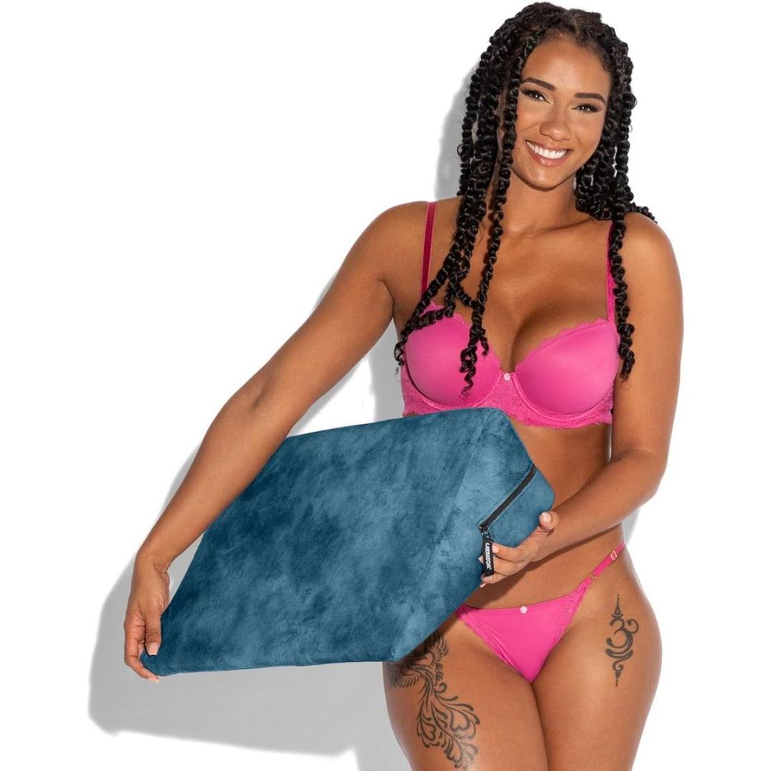 A woman in bright pink lingerie smiles while holding the Liberator Wedge Ibiza Collection - Ocean Blue by Liberator. She has long curly hair, visible thigh tattoos, and stands against a plain white background.