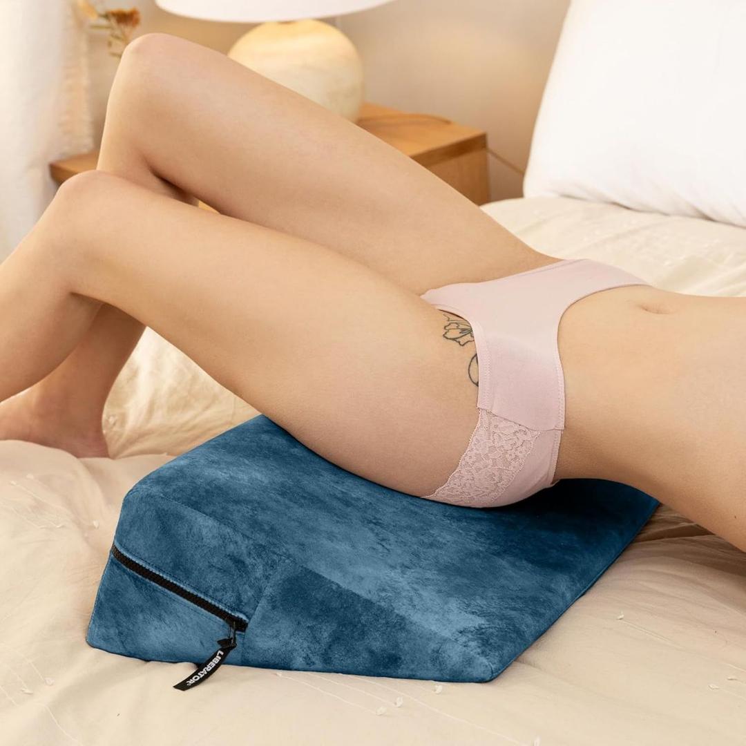 A person lies on their back with hips supported by the Liberator Wedge Ibiza Collection - Ocean Blue from Liberator, legs bent and feet flat on the bed. A bedside table and lamp are visible in the background.