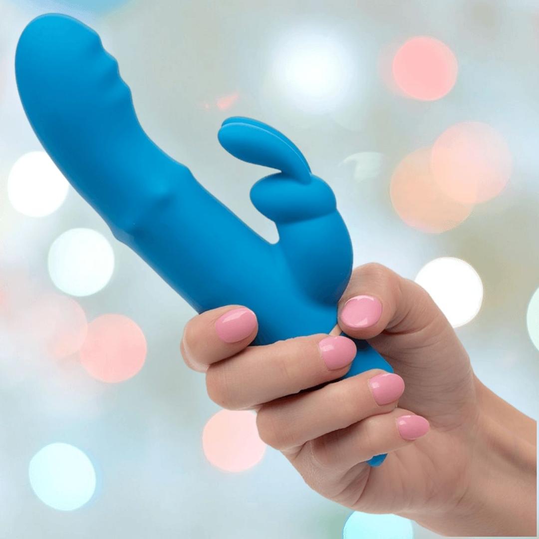 A hand with pink-painted nails holds the CalExotics Love Bunny Beaded “G” Bunny, a blue silicone rabbit vibrator, against a soft, out-of-focus background with pastel bokeh lights.