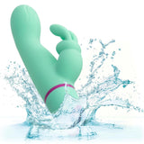 The CalExotics Love Bunny Rockin’ “G” Bunny, a light green waterproof vibrator with a curved shaft and dual stimulation arms, splashes into clear water, sending droplets flying in all directions.