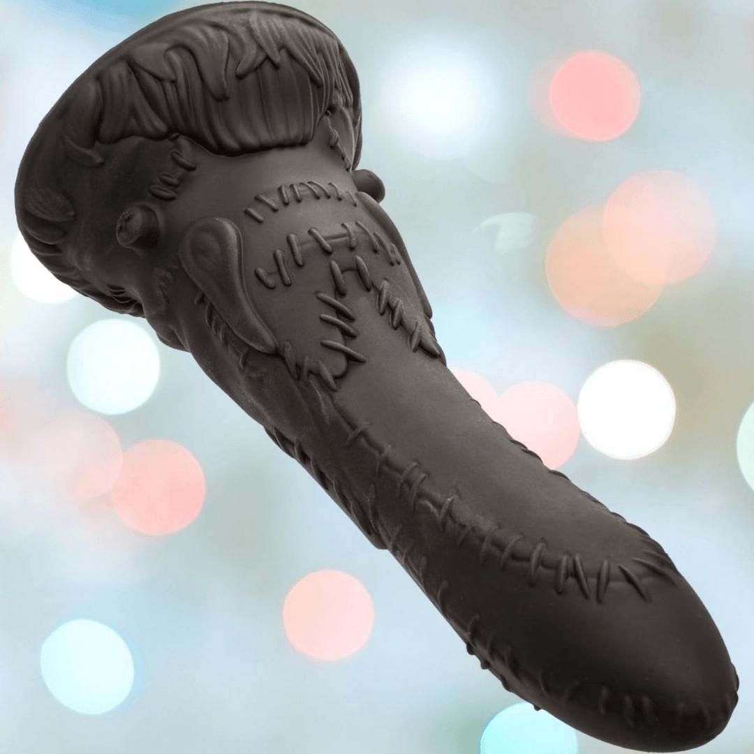 The CalExotics Monster Dong Frankenstein’s Monster is a black, sculpted premium liquid silicone toy with bulbous details, resembling a stitched monster tentacle or head, shown against blurred colorful lights.