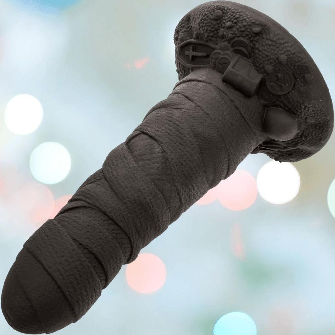 A close-up of CalExotics’ Monster Dong The Mummy, a black, textured liquid silicone dildo with a rounded tip and detailed base. The softly blurred background features pastel-colored lights.