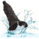 The Monster Dong The Werewolf by CalExotics, a black textured liquid silicone dildo with a flared suction cup base, is shown partially submerged in splashing water to highlight its waterproof design.