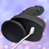 The BMS Naked Addiction Silicone Rotating & Thrusting Dildo - Noir 8.6" is displayed on a blurred purple and pink background, with a white charging cable connected to its base.