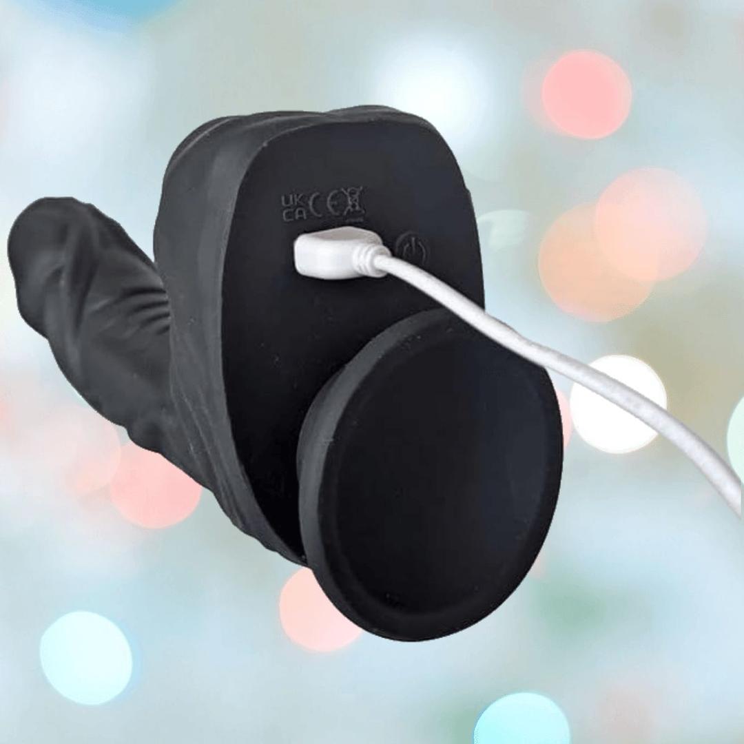 The BMS Naked Addiction Silicone Rotating & Thrusting Dildo-Noir 8.6" is displayed charging with a white USB cable, its realistic black IncrediFeel Silicone texture shown against a soft pastel background.