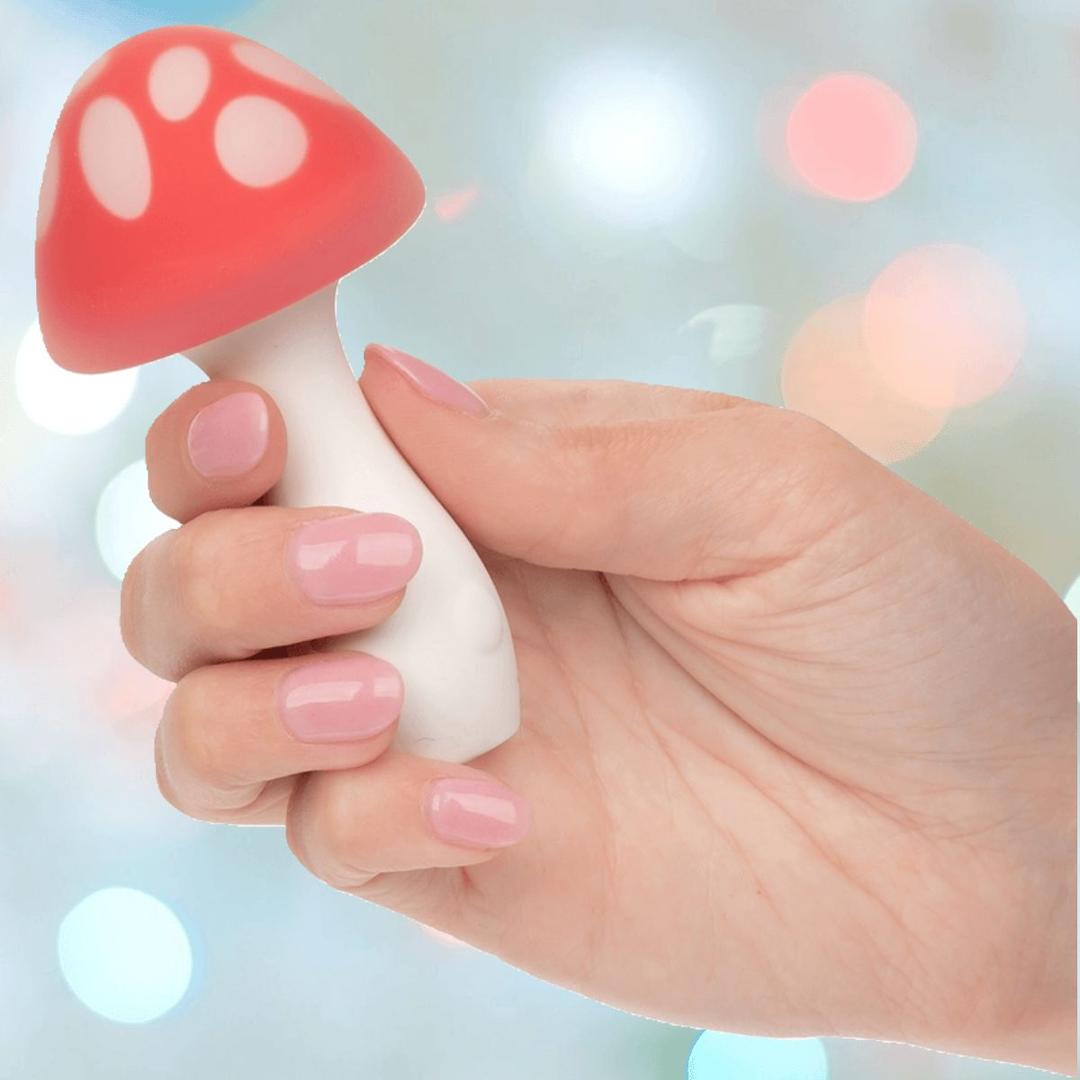 A hand with light pink nails holds the CalExotics Naughty Bits Muff Shroom Playful Massager, a small toy with a white stem and a red cap featuring white spots. The softly blurred background features pastel-colored lights.