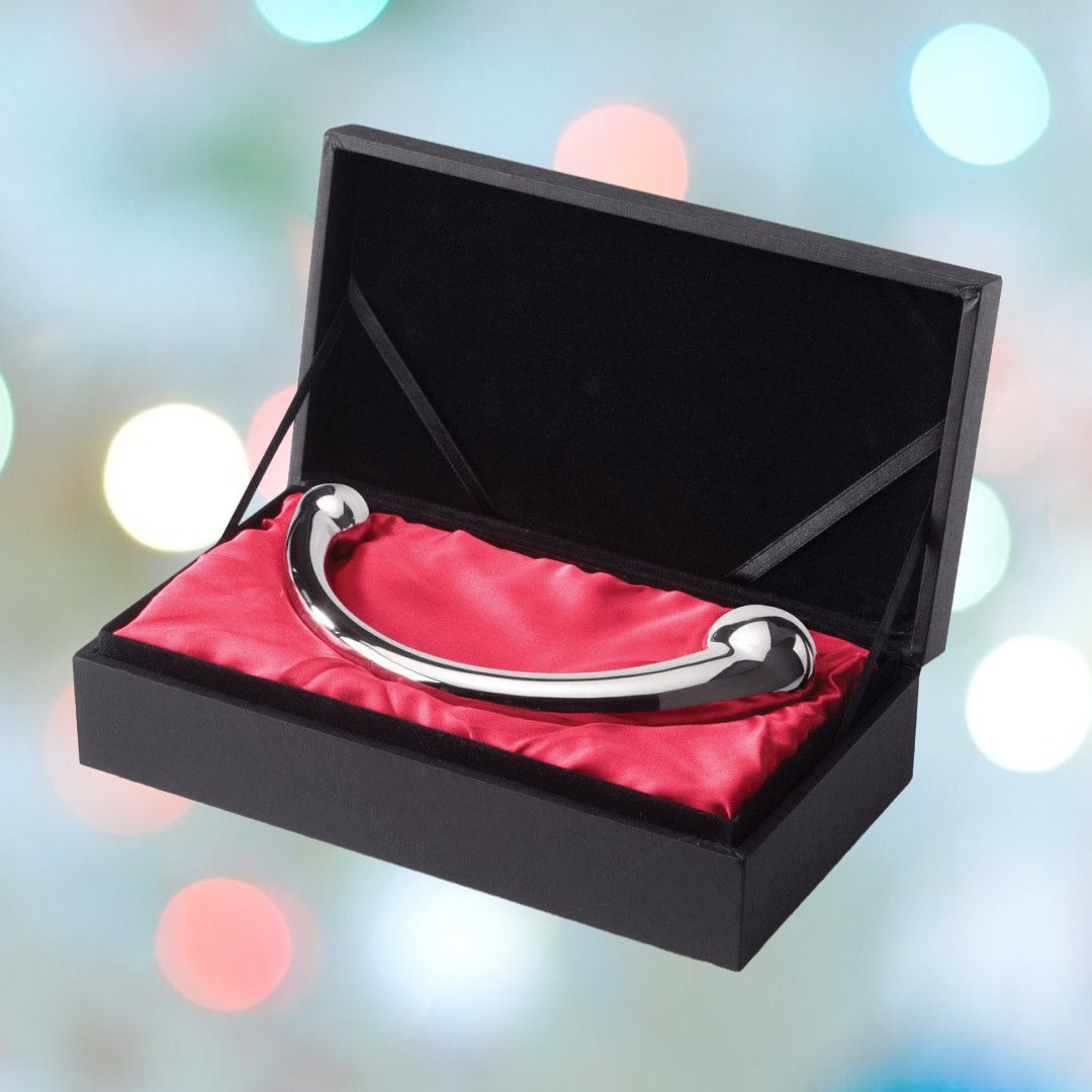 A black velvet box with a red satin interior cradles the shiny, flawless curvature of the Njoy njoy Pure Wand Non-Vibrating Metal G-Spot and P-Spot Massager, crafted from medical-grade stainless steel. The background is softly blurred with colorful bokeh lights.