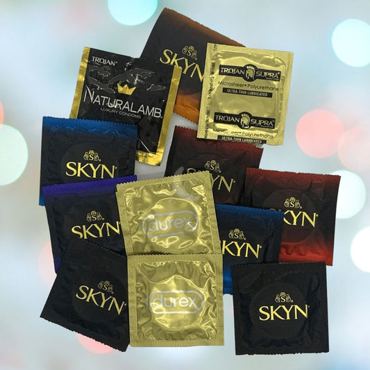 Various packages of the Condomania Non-Latex Condom Sampler are displayed, showcasing brands such as SKYN, Durex, Trojan Supra, and Naturalamb. These packages feature non-latex condoms in black, gold, and yellow tones and are arranged against a light abstract background. 1080