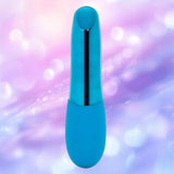The Nu Sensuelle Nikita by NOVEL CREATIONS USA, a smooth deep turquoise cylindrical bullet vibe with a rounded top, is displayed against a softly blurred pastel purple and pink background.