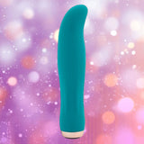 The Nu Sensuelle Velvet Touch Bella by NOVEL CREATIONS USA in emerald green stands upright against a blurred background with pink, purple, and orange bokeh lights, crafted from velvet touch liquid silicone for luxurious sensations.