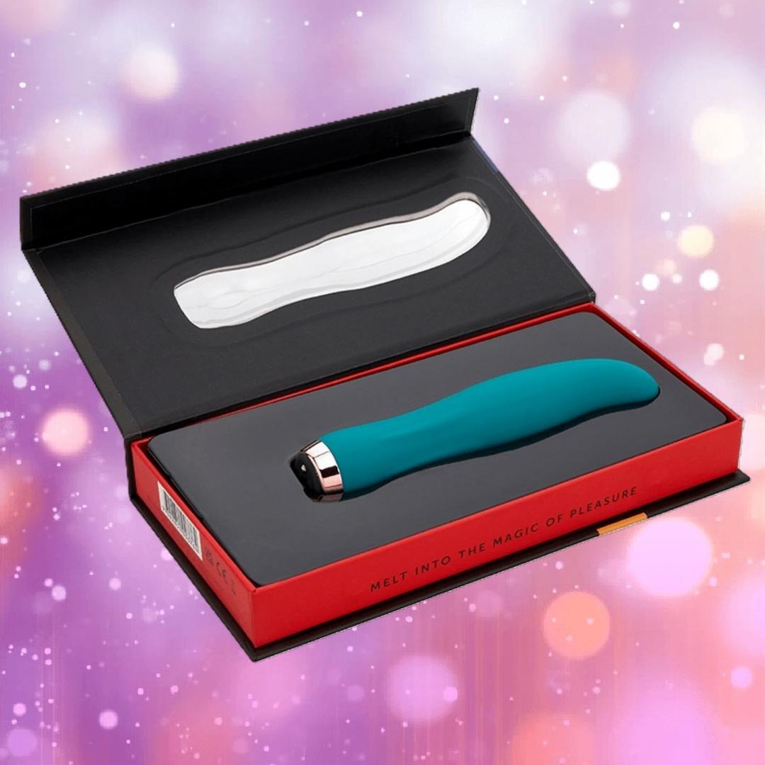 An emerald green Nu Sensuelle Velvet Touch Bella by NOVEL CREATIONS USA sits in an open red and black box with “Melt into the magic of pleasure,” against a soft background of pink and purple bokeh lights.