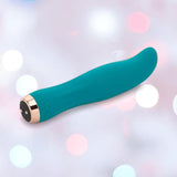 The Nu Sensuelle Velvet Touch Bella Vibrator with Turbo Boost by NOVEL CREATIONS USA features an emerald green, curved design and gold base made from body-safe silicone, shown against a dreamy pastel-lit backdrop.