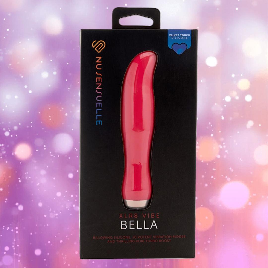 The image features a boxed Nu Sensuelle Velvet Touch Bella in hot pink by NOVEL CREATIONS USA, with a heart icon for velvet touch silicone. The background is purple with blurred light spots and sparkles.