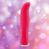 The Nu Sensuelle Velvet Touch Bella vibrator by NOVEL CREATIONS USA, in hot pink with a smooth velvet silicone surface and curved tip, stands upright against a blurred purple and peach bokeh background.