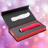 The Nu Sensuelle Velvet Touch Bella - Hot Pink by NOVEL CREATIONS USA, a red curved tip vibrator with velvet-touch silicone, is displayed in an open black and red box against a blurred pink and purple background with light circles.