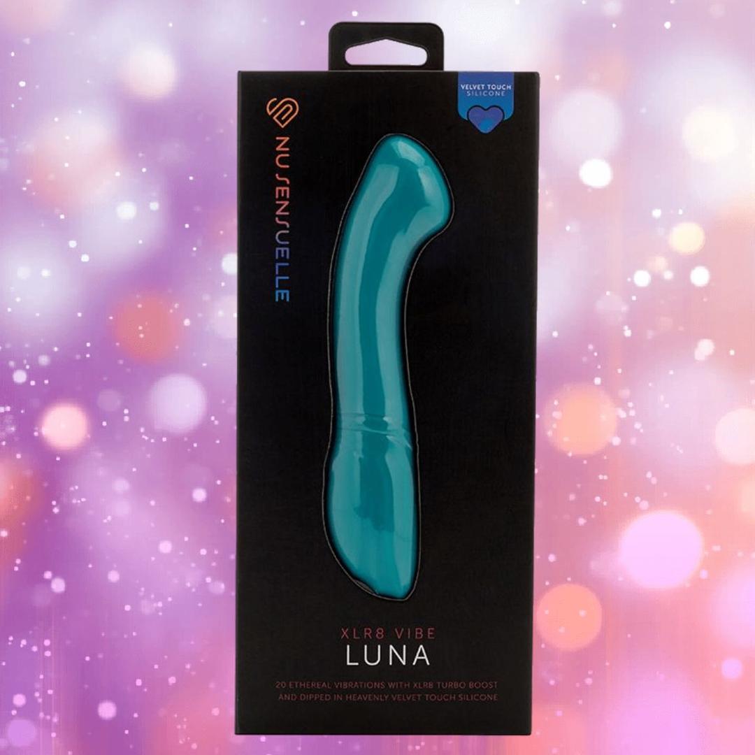 An emerald green Nu Sensuelle Velvet Touch Luna vibrator by NOVEL CREATIONS USA, featuring velvet touch silicone and 20 vibration patterns, is shown in black packaging against a pink and purple bokeh background.