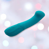 The Nu Sensuelle Velvet Touch Luna Vibrator by NOVEL CREATIONS USA features an emerald green, curved silicone design with control buttons and 20 vibration patterns, shown against a softly blurred white and pink light background.