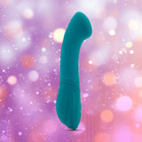 The Nu Sensuelle Velvet Touch Luna in emerald green by NOVEL CREATIONS USA stands upright against a blurred, colorful bokeh background. With 20 vibration patterns and water-friendly design, it elevates your pleasure experience.