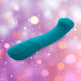 The Nu Sensuelle Velvet Touch Luna in emerald green by NOVEL CREATIONS USA, featuring a curved shape and control buttons, crafted from velvet touch silicone, rests against a shimmering pink and purple bokeh background.