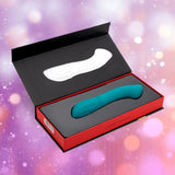 The Nu Sensuelle Velvet Touch Luna in emerald green by Novel Creations USA, featuring 20 vibration patterns and velvet touch silicone, is shown in a red and black box against a blurred pink, purple, and orange bokeh background.