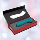 The Nu Sensuelle Velvet Touch Luna Vibrator with Turbo Boost by NOVEL CREATIONS USA, in emerald green silicone, sits in a red and black box with a white insert against a softly blurred backdrop of pink, blue, and white lights.