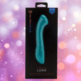 An emerald green Nu Sensuelle Velvet Touch Luna vibrator by NOVEL CREATIONS USA, featuring velvet touch silicone and 20 vibration patterns, is shown in black packaging against a pink and purple bokeh background.