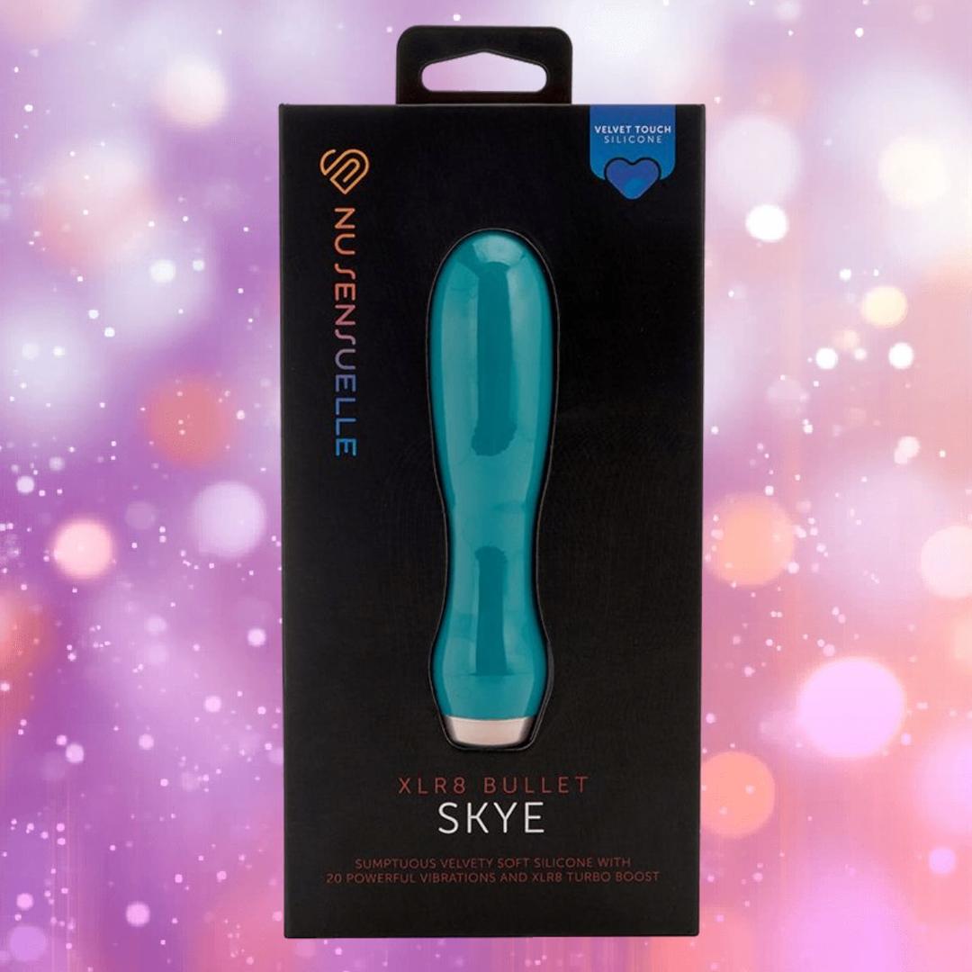 An emerald green Nu Sensuelle Velvet Touch Skye bullet vibrator by NOVEL CREATIONS USA in sleek black packaging with a purple and pink bokeh background. The box highlights "velvet touch silicone," "20 powerful vibrations," and the XLR8 turbo boost.