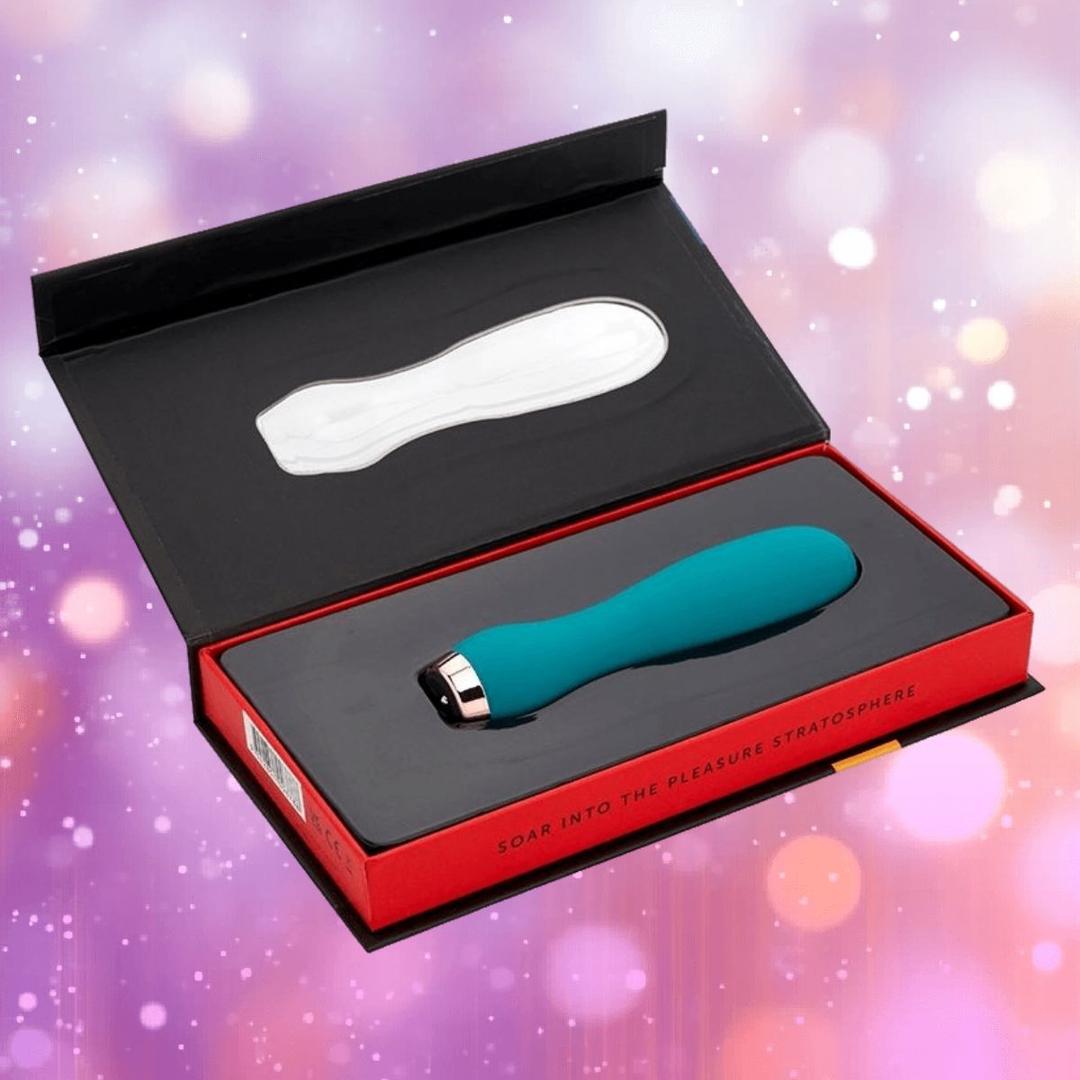The Nu Sensuelle Velvet Touch Skye by NOVEL CREATIONS USA in emerald green is displayed in its black and red box, open to reveal the vibrator against a blurred pink and purple backdrop with soft light spots.