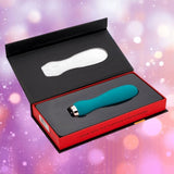 The Nu Sensuelle Velvet Touch Skye by NOVEL CREATIONS USA in emerald green is displayed in its black and red box, open to reveal the vibrator against a blurred pink and purple backdrop with soft light spots.