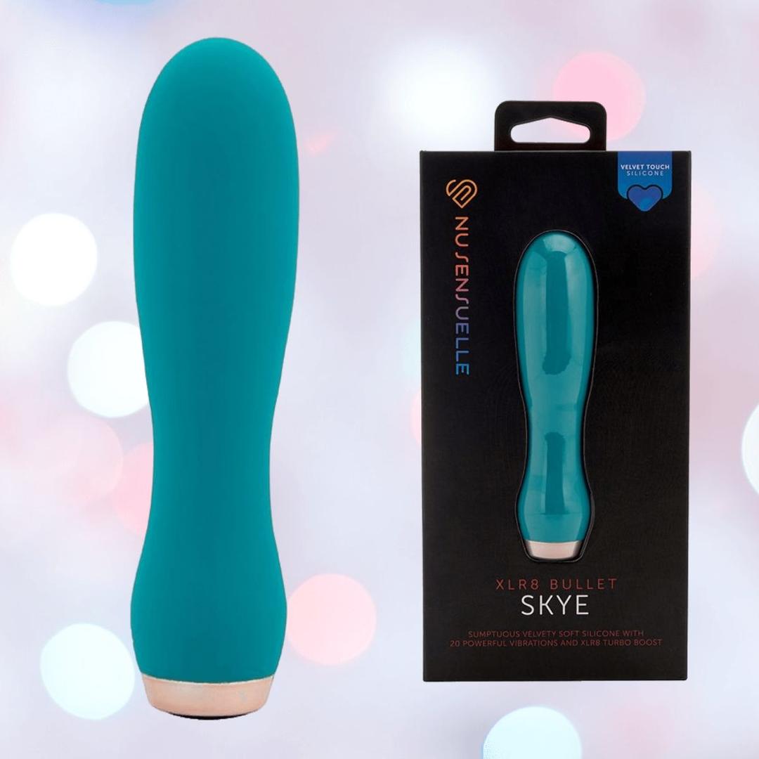 An emerald green Nu Sensuelle Velvet Touch Skye Vibrator with Turbo Boost by NOVEL CREATIONS USA stands upright next to its black box, set against a soft bokeh background. It offers 20 vibration settings for versatile pleasure.