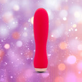The Nu Sensuelle Velvet Touch Skye by NOVEL CREATIONS USA is a hot pink, cylindrical bullet vibrator with velvet-touch silicone, shown standing upright before a backdrop of purple and peach bokeh lights.