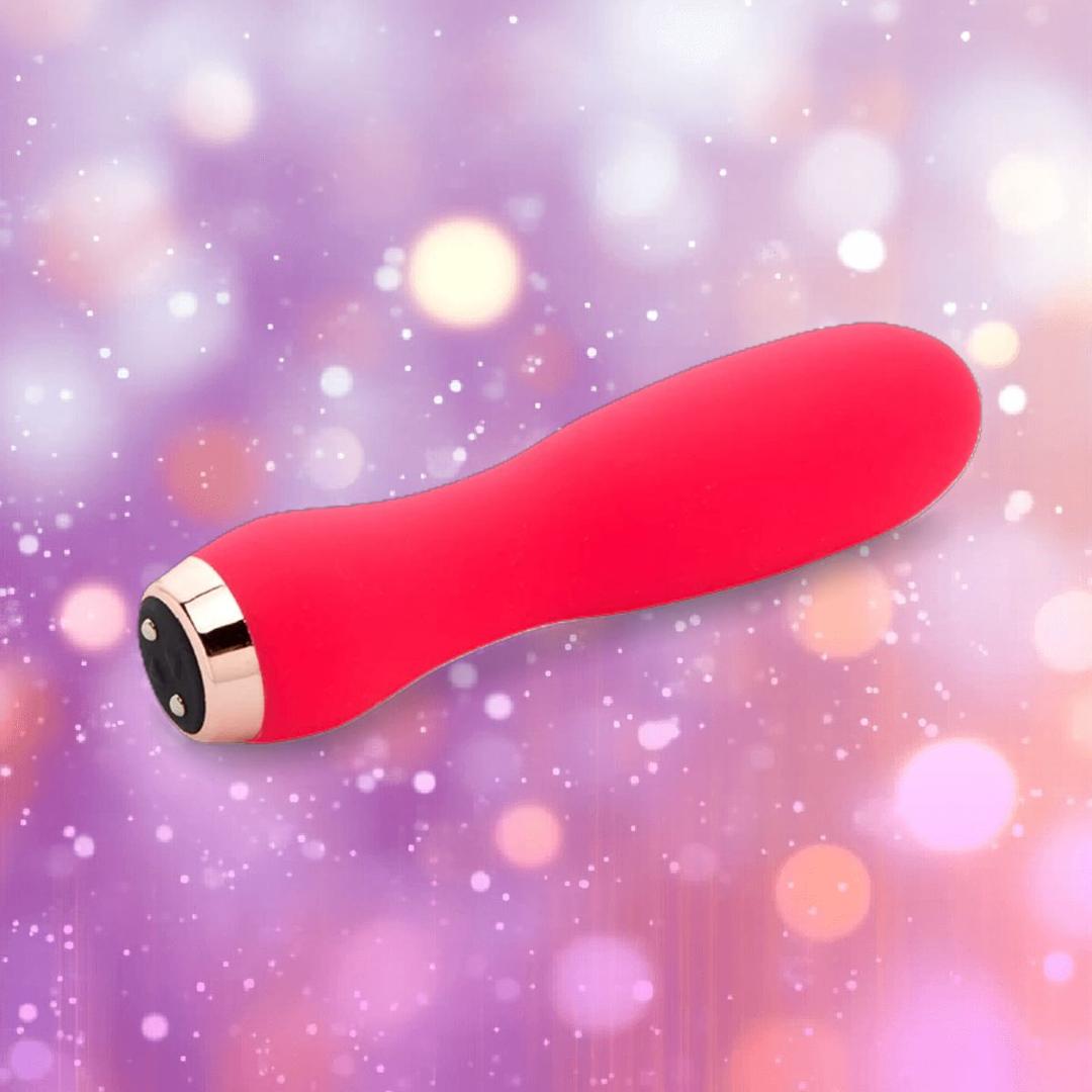 The Nu Sensuelle Velvet Touch Skye in hot pink by NOVEL CREATIONS USA is a velvet-touch silicone bullet vibrator with a rounded tip and metallic base accent, shown against a soft-focus purple and pink bokeh background.