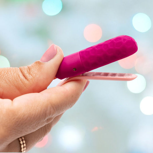 A manicured hand holds the OhMiBod Sphinx Bluetooth App-controlled Nipple Clamps in raspberry—a pink and purple textured device from OhMiBod—set against a vibrant, blurred background. 1080